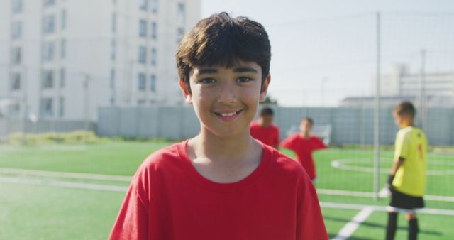 Smiling boy wearing red sports jersey standing on soccer field with teammates in background. Sunlit outdoor soccer practice featuring youth players. Perfect for topics on youth sports, teamwork, outdoor activities, or community events.
