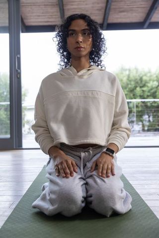 Non-binary person kneeling on mat practicing meditation, wearing hoodie and smartwatch