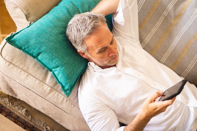 Middle-aged Caucasian man lying on a couch, using a smartphone in a cozy living room. Ideal for depicting relaxation, home lifestyle, technology use, and leisure activities. Suitable for articles or advertisements related to home comfort, technology, or lifestyle.