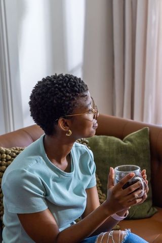 Woman relaxing on leather sofa sipping from clear mug by sunlit window in cozy living room