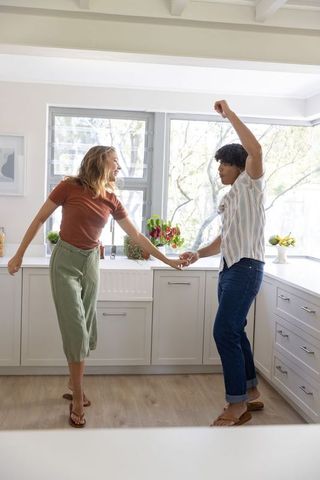 Young couple dancing in sunlit kitchen holding hands, playful romantic morning lifestyle