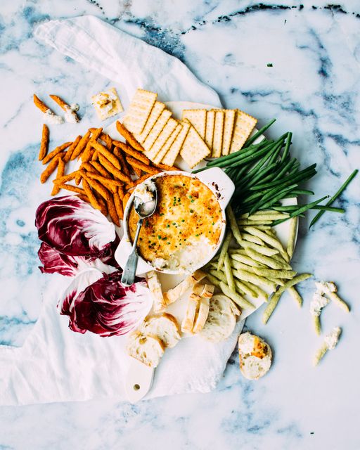 This image shows an appealing arrangement of a snack platter on a marble table. The platter includes fresh green beans, radicchio, crisp crackers, bread slices, and a creamy cheese dip. The colorful and diverse selection is perfect for illustrating healthy eating, party food ideas, or food presentation concepts. It is suitable for use in cooking blogs, food magazines, and health and lifestyle websites.