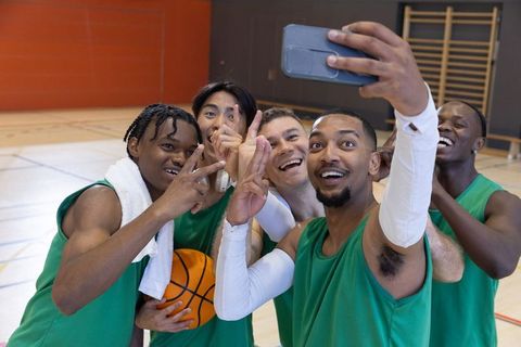 Diverse basketball teammates taking energetic selfie celebrating win on indoor court