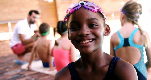 Group of children attending swimming class outdoors. Happy girl in the foreground wearing swim goggles and swimsuit, smiling at the camera. Other children are sitting on the pool deck with their instructor in the background. Ideal for use in educational, recreational, or children's activities contexts, such as flyers for swim lessons or social media posts promoting healthy lifestyle activities for kids.