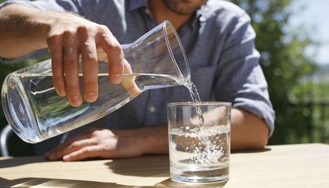 Man pouring water from glass carafe into tumbler on sunny outdoor wooden table