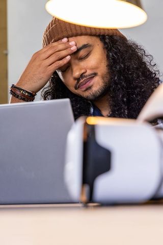 Young man rubbing forehead at desk with laptop and vr headset showing fatigue