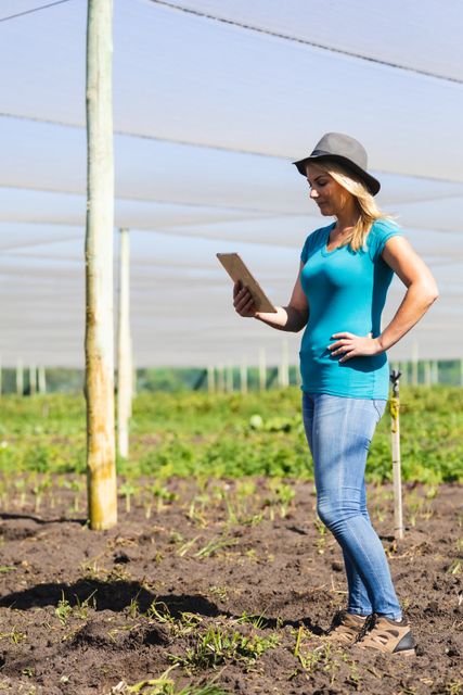 Caucasian mid adult female agronomist wearing hat using digital tablet while standing in greenhouse. summer, nature, unaltered, agronomist, research, technology, organic farm and farming concept.