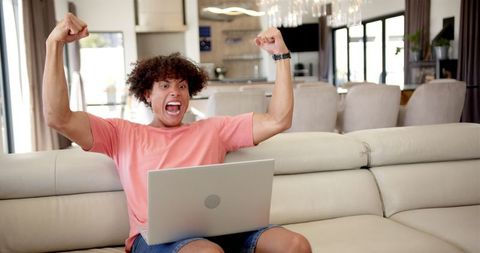 Young man with curly hair and pink shirt celebrating a victory while using a laptop on a beige sofa at home. Perfect for illustrating concepts of winning, success in online activities or work, and joyful reactions to digital content. Useful for articles, marketing materials, or blog posts focusing on technology, excitement, and positive outcomes.
