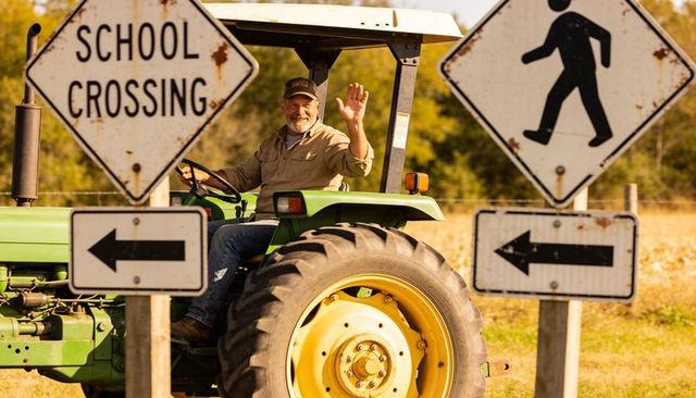 Waving farmer driving green tractor past school crossing signs on sunlit rural road