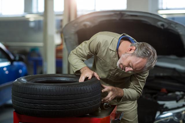 Mechanic in uniform examining a tire in an auto repair garage. Ideal for use in content related to automotive repair, vehicle maintenance, professional services, and car workshops. Can be used in articles, advertisements, and websites focusing on car repair and maintenance services.