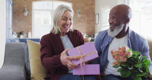 Happy senior diverse couple in living room sitting on sofa, giving flowers and present. retirement lifestyle, spending time at home.