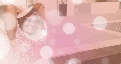 Sunlit senior woman adjusting straw hat by poolside deck with dreamy bokeh light