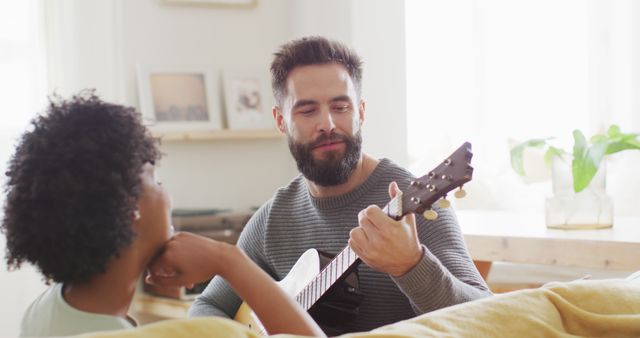 Image of rear view of happy diverse couple at home, man playing guitar, singing to woman, copy space. Happiness, inclusivity, free time, romance, music, togetherness and domestic life.