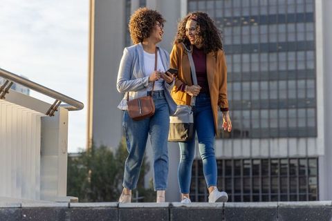 Walking and laughing african american women on city walkway holding smartphone and crossbody bags