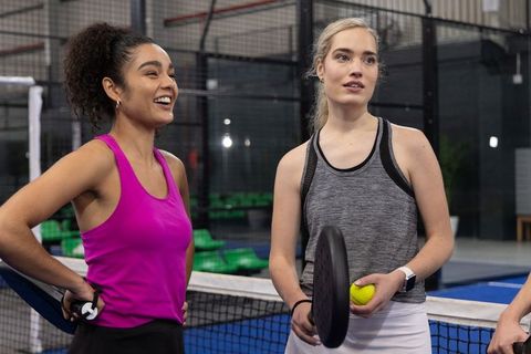Two women playing pickleball on indoor court holding paddles and yellow ball in activewear