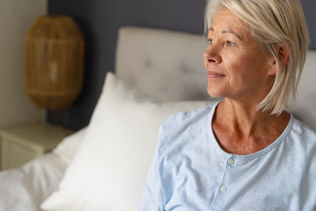 Smiling mature caucasian woman sitting on bed looking away towards window, copy space. Domestic life, living alone and senior lifestyle concept.