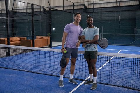 Smiling diverse men posing at indoor padel net on blue court holding rackets and ball