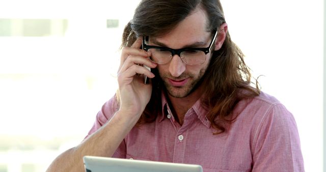 This image depicts a young professional man with long hair and glasses talking on the phone while looking at a tablet. He is wearing a casual pink shirt and appears to be working in a modern office environment. This photo can be used in contexts related to business, technology, remote work, and communication.