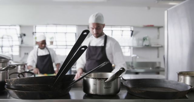 Chefs in uniform are working in a commercial kitchen. Focused on the foreground, there are several cooking utensils and pans on a counter. This image can be used to illustrate topics related to the culinary industry, commercial cooking environments, and professional kitchen settings.