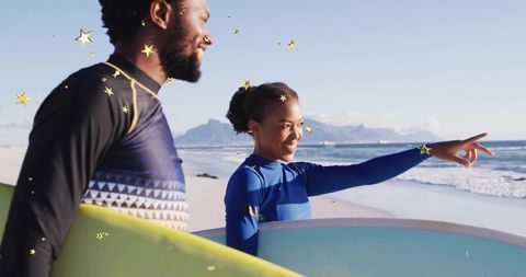 Couple On Beach Pointing At Ocean Wearing Wetsuits Holding Surfboards
