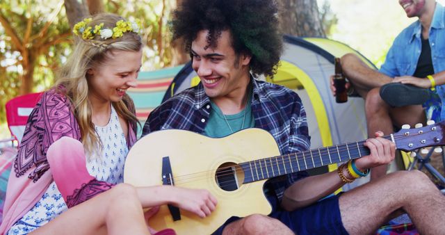 Young diverse friends are enjoying a camping trip filled with laughter and music. A young man is playing the guitar while a young woman smiles and interacts with him. They are surrounded by trees and a tent, indicating a fun and relaxing outdoor experience. This image is perfect for promoting outdoor activities, summertime fun, group outings, music festivals, and leisure experiences.