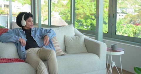 Mid-adult man relaxing on sofa with headphones in sunlit modern minimalist living room