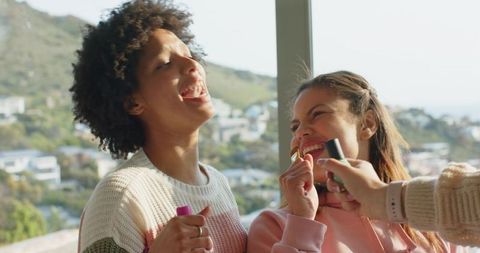 Diverse friends laughing while applying lipstick by sunlit window