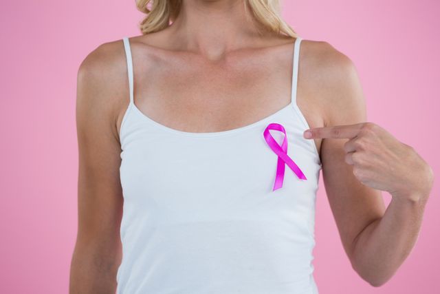 Mid section of woman pointing on Breast Cancer Awareness ribbon while standing against pink background