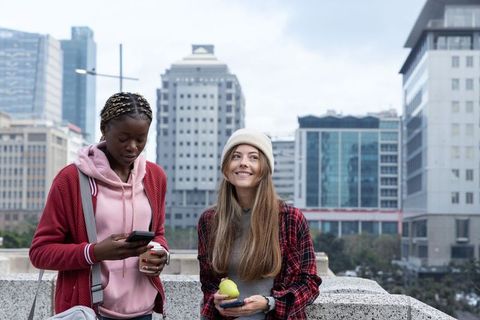 Diverse young women enjoying urban terrace using smartphones holding coffee and apple snack