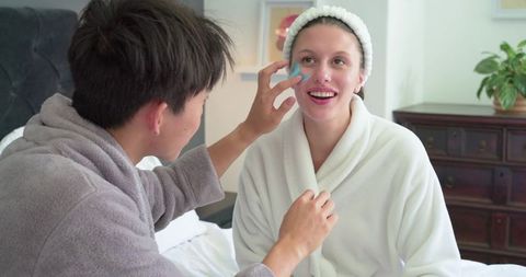 Diverse couple sharing skincare routine in bedroom robes applying blue facial sponge