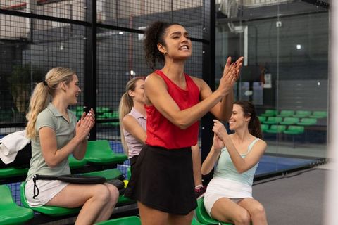 Diverse female teammates cheering on indoor glass court with vibrant green seating