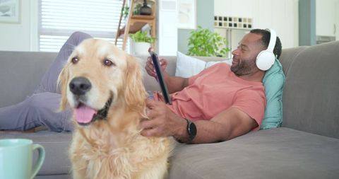 African american man reclining on sofa listening with headphones petting golden retriever