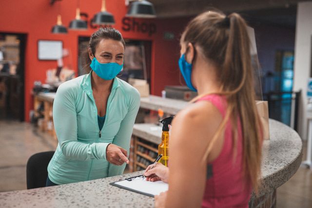 Two women wearing face masks are talking at the reception of a gym. One woman is filling in a form while the other is pointing at it. This image can be used to depict health and safety measures in fitness centers during the COVID-19 pandemic. It is suitable for articles, blogs, or promotional materials related to gym safety protocols, fitness during the pandemic, or general health and wellness topics.