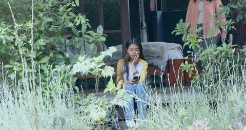 Asian woman sitting on wooden porch steps scrolling smartphone among garden plants