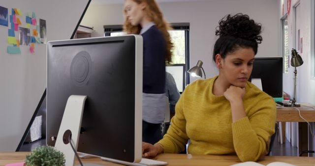 Professional woman in yellow sweater concentrating at computer screen in a modern office with coworker walking by. Suited for business, technology, and office environment themes. Useful for depicting modern workplace settings and female professionals at work.
