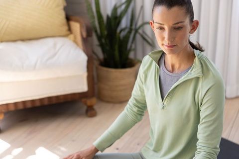 Calm woman sitting on yoga mat practicing mindfulness at home in pale green activewear