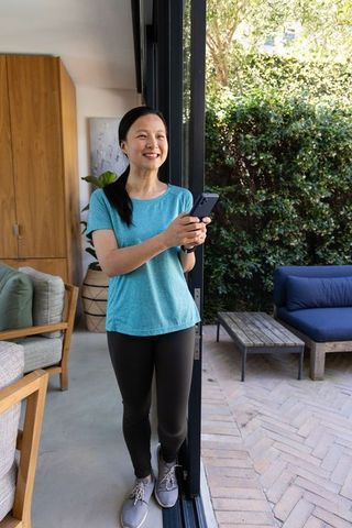 Smiling asian woman standing in sliding-door threshold holding smartphone on sunlit patio