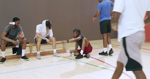 Diverse basketball teammates taking break hydrating on bench after indoor training session
