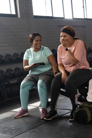 African american women laughing and sharing smartphone on weight bench in gym