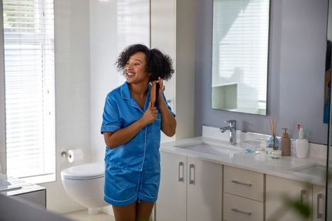 African american woman in blue pajamas grooming hair in modern bathroom