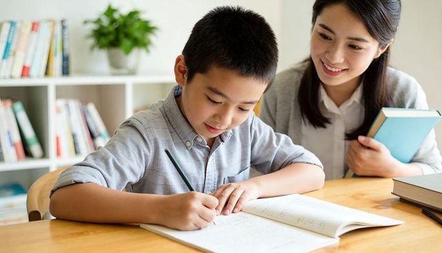 Young student writing workbook while tutor smiling holding book during home study