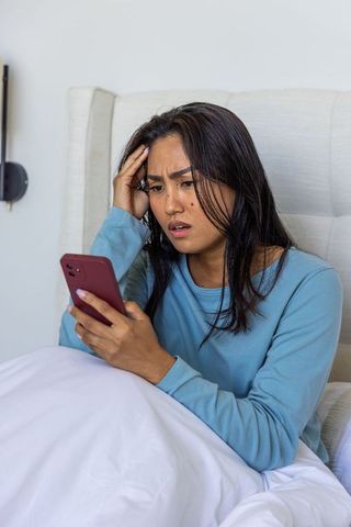 Asian woman reacting with shock while holding smartphone in bed, modern loungewear