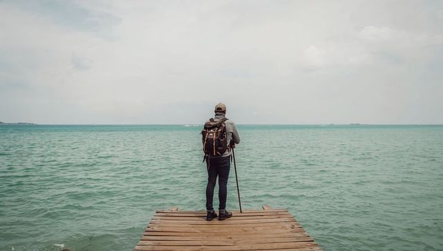 Hiker standing on wooden dock facing turquoise sea with backpack and walking stick