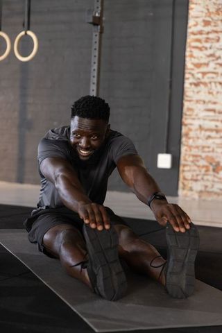 African american man stretching hamstrings on mat in gym smiling while wearing smartwatch
