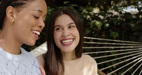 Diverse women laughing on hammock in backyard garden wearing knit sweaters and gold hoops