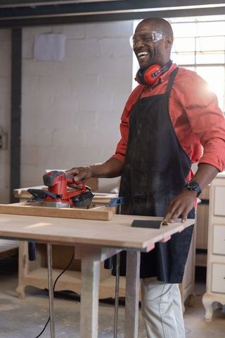 African american carpenter sanding wood plank in shop wearing goggles and ear protection