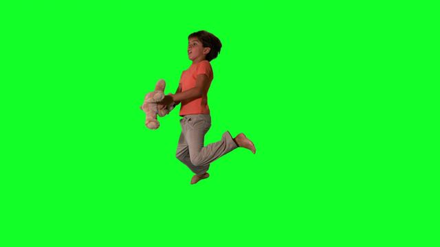 Boy in bright shirt jumping joyfully and holding a teddy bear against a green screen background, ideal for children-related projects or education visuals. Can be used for state-of-the-art banners, playful flyers, or energetic marketing content requiring an element of innocence and cheer. Use of green screen allows versatile alteration for creative backgrounds.