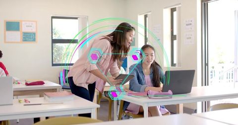 Instructor guiding student using laptop and tablet in modern classroom with digital overlay