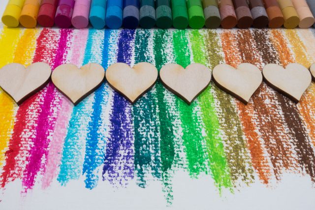 Vibrant and visually appealing composition of wooden hearts placed on top of colorful pastel crayons with rainbow stripes in the background. Perfect for art and craft-related projects, Valentine's Day themes, wedding invitations, or any creative design work emphasizing creativity and love.