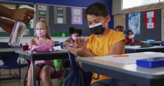 Biracial schoolboy wearing face mask disinfecting hands in classroom, colleagues in background. children in primary school during coronavirus covid 19 pandemic.